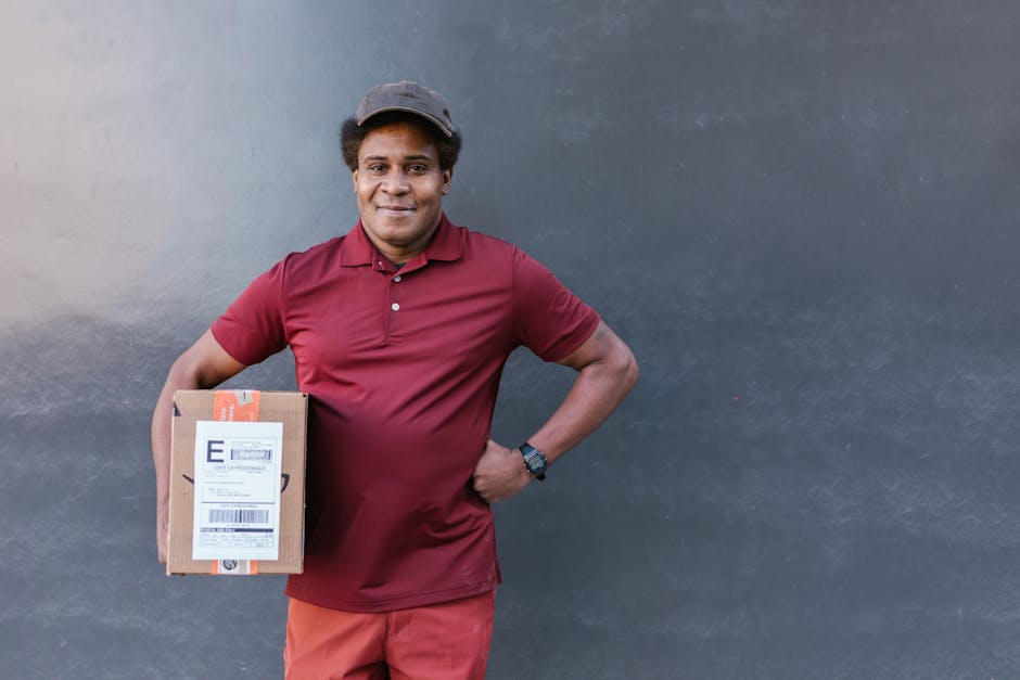A smiling male delivery person with dark curly hair, wearing a maroon polo shirt, beige cap, and watch, stands against a plain, dark grey wall. He holds a small cardboard parcel with a label and orang