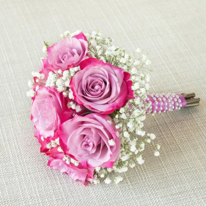 An enchanting wedding bouquet from the heart of Stanmore blends romance with a contemporary flair, perfect for the local bride or special celebration. At its core, five radiant roses command attention, their petals painted by nature in gradients of rich fuchsia pink and calming lavender. The edge of every petal deepens into magenta, creating an effect of depth and movement. Dainty sprigs of snow-white baby's breath nestle between these roses, infusing the bouquet with a soft volume and a frothy, ethereal look reminiscent of fresh morning walks in Bentley Priory Nature Reserve. The tightly bound, short stem handle is encased in sumptuous, deep plum ribbon, elegantly wound with spiral rows of luminous pearls for an added note of sophistication and tactile interest. The exposed, neatly trimmed stems anchor the bouquet in natural beauty. Placed upon a creamy, textured linen surface, the bouquet's vibrant palette gleams beneath softly diffused lighting, its details highlighted as though ready to be carried down the aisle at St John's Church or set at a family celebration in Stanmore. This thoughtful design is suited for bridesmaids, flower girls, or as a cherished centrepiece, encapsulating the blend of local tradition and modern artistry unique to Stanmore.