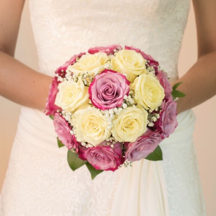 This evocative image, crafted by a skilled Stanmore florist, offers a close, personal view of a bride's embrace: her arms curve protectively around a lavish, rounded bouquet filled with locally sourced blooms. At the bouquet's heart are roses in gentle lemon yellow-fresh as the morning light that filters through Stanmore's quiet residential streets-surrounded by vibrant fuchsia and mauve-pink roses, their hues capturing both modern flair and timeless romance. Sprigs of exquisite baby's breath add a light, frothy touch among the plush petals, while deep green leaves peek from the sides, anchoring the burst of color. The bride's gown, visible in the upper frame, boasts delicate lacework reminiscent of Stanmore Hall's ornamental designs, falling softly into a flowing skirt. The image is flooded with soft, even light, illuminating every nuance in the floral arrangement and the dress's intricate textures. The serenely blurred background suggests a private moment amidst Stanmore's leafy avenues. This bouquet, shaped with devotion and expertise, captures the spirit of a wedding in Stanmore-memorable, elegant, and brimming with heartfelt local craftsmanship.