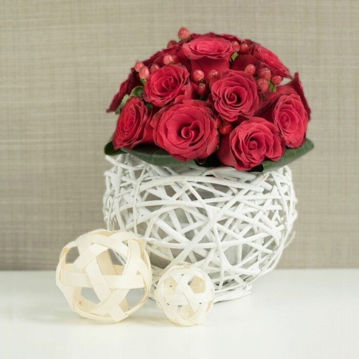 On a bright white tabletop, a dramatic arrangement of red roses and berries captures the eye, evoking the sense of an elegant wedding or anniversary dinner set somewhere in Stanmore. The central bouquet forms a dense, dome-shaped cluster of deep crimson roses, each bloom layered with silky petals that spiral outwards in rich, velvety folds. The roses are placed so closely that their petals overlap, creating a plush and continuous surface that rises confidently above the rim of the container. Between the rose heads, small sprays of glossy, deep red hypericum berries are tucked in, adding polished highlights and a slightly varied shade of red, like tiny jewels nestled in the blooms. A low collar of dark green foliage supports the bouquet, mostly hidden but offering the faintest glimpse of leaf texture beneath the sea of red. This opulent floral dome emerges from a striking spherical vase crafted from white-painted branches or wicker. The twigs are arranged in an intricate open lattice, forming a hollow globe that feels light and airy despite its sculptural presence, allowing the viewer to see through the gaps and notice the play of light and shadow on the stems within. Resting to the left on the same clear white surface are two decorative wicker balls in natural beige or cream tones. One is noticeably larger, placed nearer the front, while the smaller sphere sits slightly behind, their loose basket-weave pattern subtly mirroring the vase but in warmer, more organic hues that would complement a modern Stanmore interior. In the background, a softly blurred wall in pale taupe or beige with a faintly textured finish recedes gently, keeping the focus on the flowers. The entire composition feels refined and romantic, perfect for a top table centerpiece or intimate dinner arrangement, easily imagined arriving fresh from a skilled florist serving Stanmore.