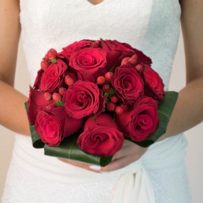 This finely detailed image captures a bride in Stanmore, visible from the waist up and resplendent in a bright white wedding dress adorned with floral lace overlay and delicate V-neck, gently supporting a truly opulent bridal bouquet. Illuminated by soft, even daylight, the star of the composition is a lush, compact sphere of velvety red roses-each rosette tightly coiled or just beginning to open, pressed close for a plush, almost intertwined effect. Among the blooms are spirited splashes of glossy bright red hypericum berries, whose rounded forms peek out with cheerful vibrancy, infusing the bouquet with depth and visual delight. The base is meticulously finished with broad, deep green tropical leaves, hand-folded and wrapped into a structured, glossy collar that gives the entire arrangement presence and polish. The bride's hands cradle the bouquet tenderly, manicured nails echoing the bouquet's refinement, and the skirt of her dress softly pools to one side. Just beyond, the background fades to a gentle blur-perhaps hinting at the subtle interiors of Stanmore's own wedding venues-ensuring all attention rests on this stunning symbol of love, passion, and bespoke skill. It's a bouquet designed for lasting impression, the sort often requested for unforgettable ceremonies at Chandos Park or Stanmore Golf Club, reflecting both floral tradition and local expertise.