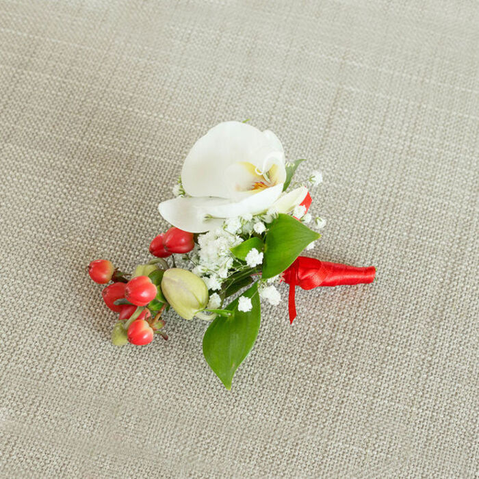Displayed against a softly textured, oatmeal-toned linen background, this elegant boutonniere appears as though it has just been set down on a dressing table before a ceremony at a local Stanmore venue. The fabric's natural weave creates a gentle backdrop that lets every detail of the floral work stand out clearly. At the heart of the piece blooms a pristine white Phalaenopsis orchid, its generous petals unfolding like porcelain wings, with a subtle flush of creamy yellow and soft orange deep in the throat that adds both depth and warmth. Surrounding this central orchid, clusters of fine white baby's breath create an ethereal haze, their tiny blossoms forming a delicate, cloudlike border that softens the structure of the buttonhole. To one side, a compact spray of round, glossy red berries introduces a lively, festive contrast, their surfaces bright and reflective, while a handful of lighter green, oval buds offer a quieter, fresher tone. Interwoven through the flowers and berries are lush green leaves in varied shapes-some broader and pointed, others slender and tapering-giving the arrangement a sense of natural volume and a hint of movement, as if just gathered from a well-kept garden in Stanmore's residential streets. At the base, the stems are neatly wrapped in a vivid red satin ribbon, smooth and slightly lustrous, tied into a careful knot with short trailing ends that rest flat against the cloth, ready for pinning to a lapel. The composition conveys an impression of refined celebration, ideal for a groom, groomsman, or special guest attending a wedding, prom, or seasonal event in and around the Stanmore area.