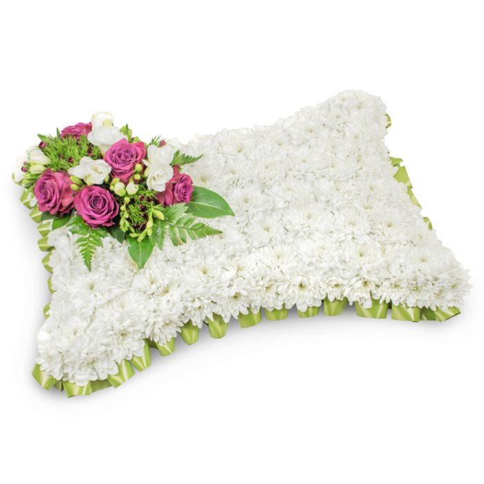 A rectangular memorial pillow, composed entirely of small, snow-white chrysanthemums, lies against a pristine white backdrop and radiates a soft, comforting simplicity. The chrysanthemums are arranged so densely that the surface appears like a plush textile - each tiny bloom contributing to a unified field that evokes serenity and rest. Positioned deliberately on the upper-left corner, a cluster of deep magenta and fuchsia roses forms the visual heart of the piece; their rich tones and sculpted petals stand in graceful contrast to the white ground. Among those roses, delicate white freesias and small white buds weave in and out, introducing an ephemeral sweetness and a refined contrast of scale. Lush green foliage - including slender fern fronds and glossy leaves - frames the accent flowers and leads the eye along subtle diagonal lines, balancing the composition with organic movement. Around the pillow's perimeter a fine pleated ribbon in pale fresh green is tucked beneath the chrysanthemums, its satiny texture visible in places and lending a quiet ceremonial edge. The photograph's neutral setting, with soft shadows beneath the form, highlights the three-dimensional craftsmanship and the arrangement's intended purpose: a dignified sympathy tribute for funerals, memorials, or a graveside farewell. With a feeling of hush and repose, this arrangement is the sort of floral cushion families in Stanmore might choose to place near a coffin or at a cemetery plot, offering a tactile, floral expression of remembrance and consolation.