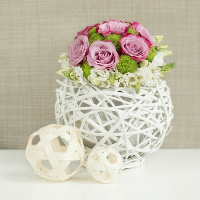 This photograph presents an elegant floral arrangement perfectly suited to a Stanmore wedding breakfast or a refined dinner party overlooking Stanmore Country Park. The focal point is a low, rounded bouquet of roses and accent blooms, shaped into a neat dome that spills just slightly over the rim of its container. The roses, in a harmonious mix of soft pink and gentle lavender, are fully opened, their petals forming plush, layered rosettes. Some petals deepen to a richer pink at the edges, giving the flowers a nuanced, romantic character. Interspersed among the roses are clusters of bright green button chrysanthemums, their compact, bobble-like heads creating a pleasing textural contrast and introducing a fresh, garden-bright note. Encircling the base of the bouquet are small, white filler flowers, which peek out between the larger blooms and lend the arrangement a delicate, airy finish. The flowers are arranged in a distinctive white vase shaped like a sphere, composed of intertwined rattan-style branches that form an open, basket-like weave. This airy structure allows glimpses of the stems inside and casts subtle shadows on the smooth white tabletop beneath, enhancing the sense of craftsmanship. To the left of the vase, two decorative woven spheres in pale, natural tones-one larger, one smaller-rest on the same surface, their open lattice pattern echoing the vase's design and tying the composition together. Behind it all, a softly blurred beige background with a gentle fabric texture keeps the visual focus firmly on the flowers while adding warmth. The image conveys calm sophistication and gentle celebration, making it an ideal representation of a wedding table centerpiece or special occasion floral design in and around Stanmore.