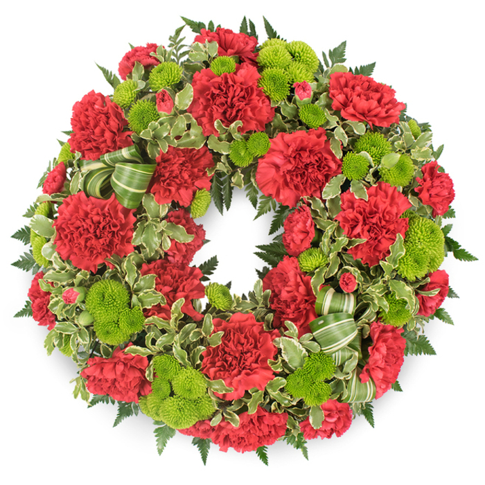 A lush, circular memorial wreath commands attention against a pure white background, its ring of flowers forming a continuous flow of deep red and bright lime green. The wreath is crafted with the care of an experienced Stanmore florist, each element placed to create a sense of balance and quiet strength. Deep red carnations dominate, the ruffled layers of each bloom overlapping like soft, velvety waves. Some blossoms are wide open, showcasing their intricate petals, while others are partially unfurled, giving subtle shifts in shade and form as your eye travels around the circle. Between these red blooms, lime green button chrysanthemums nestle snugly, their small, spherical heads and tightly coiled petals introducing a different texture and a spark of fresh colour. The contrast between the crimson and chartreuse tones adds a contemporary edge to what is otherwise a very classic funeral design. Supporting the flowers is a generous base of greenery: dark green leatherleaf fern fans out with delicate, feather-like fronds, while variegated pittosporum, with its creamy white leaf margins, adds another layer of interest and softens the edges of the wreath. A ribbon in coordinated shades of green, marked with subtle stripes, is woven thoughtfully into one section, forming modest loops that sit quietly among the foliage rather than dominating it. The bright white background and even studio-style lighting reveal every detail clearly, with a faint, soft shadow beneath the wreath hinting at its three-dimensional presence. This is the sort of respectful tribute you might choose for a funeral at a local Stanmore chapel, crematorium, or cemetery, offering a dignified expression of sympathy to families across Stanmore, Harrow Weald, and along the quiet residential roads near Stanmore Country Park.