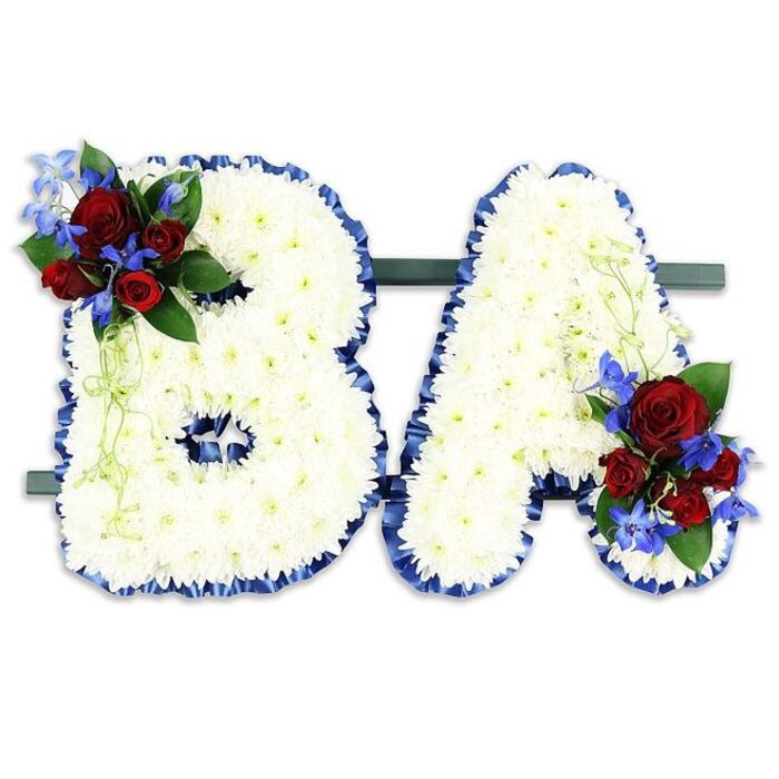 This composed funeral tribute spells the initials BA in large, upright letters, each letter densely sheathed in a cushion of pristine white chrysanthemum blooms that read as soft and luminous against a simple white background. The edges of both letters are neatly wrapped with a rich royal blue satin ribbon, its sheen and ruffled trim providing a refined border that accentuates the letterforms. On the upper left curve of the B there is a concentrated spray of deeply coloured red roses - some in tight buds, others opening to reveal velvety petals - interspersed with vertical flashes of intense blue delphinium-like flowers and set among dark green leaves; a corresponding cluster adorns the lower right of the A, with pale green trailing foliage that gently softens the corner. The two initials sit side-by-side on a slim dark green rectangular stand, giving the impression of a unified, freestanding tribute suited for placement at the front of a chapel or beside a hearse. The overall texture contrasts the matte, dense chrysanthemum heads with the glossy ribbon and the soft, tactile quality of the roses; together these elements create a composition that is at once pure, expressive and measured. I imagine this piece being chosen for memorial services in and around Stanmore, near Stanmore Tube or the leafy avenues by Bentley Priory, where families often seek a personalised emblem of remembrance. The subtle fragrance of roses and the solemn palette of white, red and blue make this an elegant and respectful floral statement for a cherished farewell.