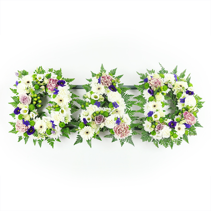In this carefully lit photograph, a bespoke funeral tribute spells out DAD in three individual floral letters, arranged from left to right against a pure white background that makes the colours appear crisp and true. Each letter is filled completely with pristine white flowers: cushion mums, daisy-style blooms, and smaller white fillers that together create a soft, almost quilted effect. Their petals overlap in gentle layers, giving a sense of comfort and peace, like a soft blanket of flowers laid in honour of a much-loved father. The outline of each letter is traced with rich green foliage, including feathery fern fronds and compact glossy leaves that form a natural border, echoing the lush greenery found around Stanmore's gardens and open spaces. Between the foliage, bright green button chrysanthemums or similar buds peep through, adding little sparks of fresh, spring-like colour. Across the letters, the florist has woven thoughtfully placed accents of purple: deep, velvety tones that resemble pansies, violas, or spray chrysanthemums, providing depth and a solemn, respectful character that feels appropriate for a funeral service. Softer purples, lavender, and pale mauve hues appear in roses and frilled carnations, their spiralling petals adding romance and warmth to the overall composition. The lighting is even and soft, as though the tribute is resting under gentle daylight in a quiet room, just before being taken to a service in Stanmore or laid at a graveside. Created with the care and precision of an experienced Stanmore florist, this DAD arrangement offers a deeply personal, traditional, and heartfelt way to say goodbye and express sympathy.