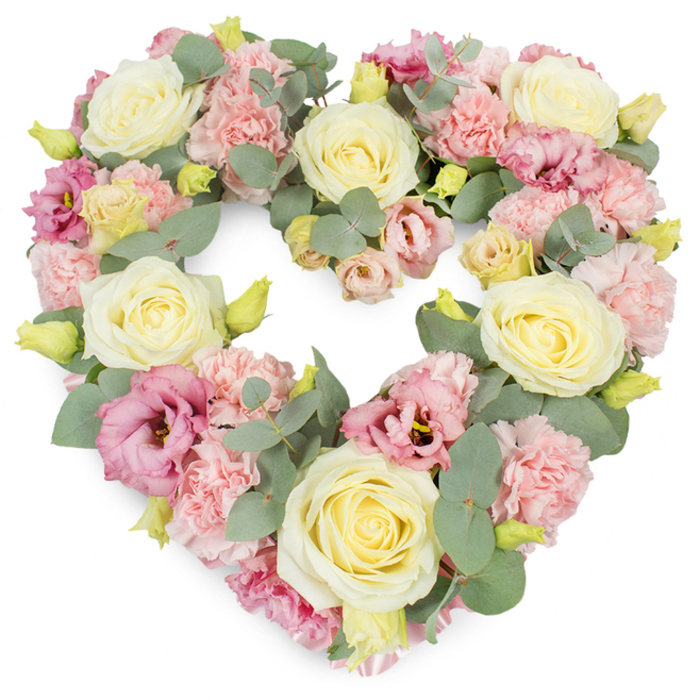 This image shows a beautifully composed, heart-shaped wreath of sympathy flowers resting against a clean white background, the design echoing the type of refined tributes often seen at services in Stanmore churches or crematoria near Canon's Park. The heart itself is flawlessly defined yet softened by its dense covering of blooms, so that the form feels both precise and tender. Cream and buttery ivory roses serve as focal points throughout, each one a full rosette of tightly layered petals, spiralling from a neat centre out to smooth, rounded edges, radiating warmth and heartfelt respect. Filling the spaces around them are generous clusters of pastel pink carnations, whose frilled, almost feathery petals overlap in multiple layers, giving the surface of the wreath a soft, pillow-like texture that invites the touch. Richer accents of colour are introduced with magenta-pink lisianthus, their slightly flared, trumpet-shaped blooms and delicate folds creating gentle pockets of shadow and depth, guiding the eye along the curves of the heart. Interwoven with this tapestry of petals are sprigs of silvery-green eucalyptus, the distinctive round leaves providing a cool, calm contrast to the warmer shades and adding a touch of natural movement. Tiny yellow-green buds can be seen peeking between petals, hinting at freshness and careful, recent assembly by an experienced florist. The overall palette of cream, blush pink, and soft green is soothing and understated, capturing a mood of peace, remembrance, and quiet gratitude. This heart-shaped floral wreath would be an especially fitting centrepiece for funeral services, memorials, or anniversary tributes in Stanmore, offering a graceful, floral expression of sympathy and enduring affection.