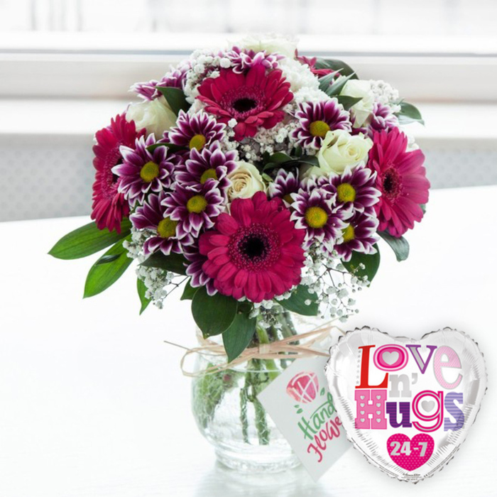 In this bright, uplifting image, a vivid bouquet and whimsical heart-shaped balloon are composed together on a clean, pale surface, as though waiting to be carried out from Flower Delivery Stanmore for a special surprise. The flowers are arranged in a stout, clear glass vase, whose transparency showcases the carefully trimmed stems standing in cool, clear water. Around the narrow neck of the vase, a piece of rustic natural twine is tied in a neat bow, supporting a simple white tag. The tag features a petite pink rose illustration and a cheerful line of green, hand-written-style text, reinforcing the impression of a thoughtfully assembled, artisan gift. The bouquet bursts into colour above the rim, forming a rounded, generous shape. At its centre, several large gerbera daisies in bold magenta-pink fan out their smooth, symmetrical petals around deep, nearly black centres, creating striking focal points that feel both fun and sophisticated. Interspersed among these are numerous smaller chrysanthemums, each bloom a two-toned marvel with dark purple petals that gently transition to bright white tips, framing sunny yellow hearts that add a touch of playful cheer. Creamy white roses are tucked throughout the design, some still partially furled, others just beginning to open, their velvety petals evoking romance and tenderness. Between the larger flowers, dainty sprays of white baby's breath (gypsophila) float like tiny stars, softening the composition, while lush green foliage peeks from all sides, grounding the design with a fresh, garden-like feel reminiscent of walks around Stanmore Country Park. To the right of the vase stands a shiny, heart-shaped foil balloon, its silver border gleaming around a white centre. The balloon is adorned with a lively mosaic of multi-coloured, patterned letters in shades of pink, purple, and blue that spell Love n' Hugs 24-7, accented with small heart icons. The softly blurred, bright background suggests a sunlit indoor setting, capturing the essence of a thoughtful flower delivery in Stanmore, perfect for expressing unwavering affection on birthdays, anniversaries, or just because.