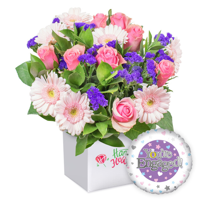 A lush, hand-tied bouquet sits in a crisp white square box, a testament to modern floristry near Stanmore. The composition centers around several full-bloom pink roses, their petals softly unfurled and resonant with romance, paired with light pink gerbera daisies that radiate cheer and clarity with white-tipped edges and pale yellow centers. Purple filler flowers peek through like bursts of evening lavender, providing rich contrast and texture, while abundant green foliage frames the arrangement with a natural, garden-fresh feel. The box's front features a delicate logo: a red rose icon beside green script, hinting at a trusted local florist. To the lower right, a round, glossy foil balloon completes the set-its purple central design encasing the words You're Engaged! in elegant silver, and a shimmering golden 'O' in You're that nods to celebration. The outer rim is sprinkled with small pastel hearts and silver stars, adding whimsy to the sophisticated display. This complete engagement gift, crafted for warmth and joy, is well-suited for congratulating couples and is available for delivery across Stanmore from Flower Delivery Stanmore, bringing brightness to any home or celebration.