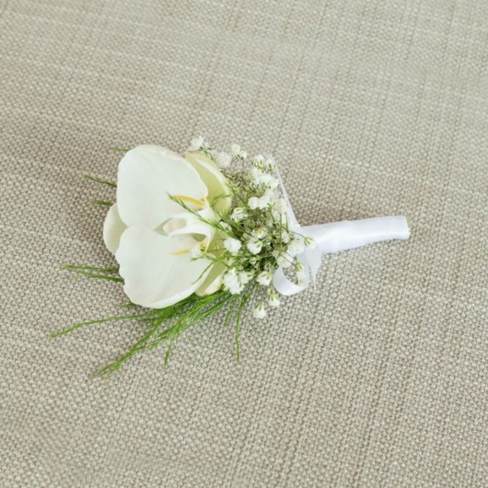 An elegant white orchid boutonniere lies horizontally across a pale beige, linen-like fabric, the gentle weave and slightly uneven texture of the background giving the scene a natural, handcrafted feel. The focal bloom is a single Phalaenopsis orchid in immaculate white, its petals broad, velvety, and slightly glossy, catching the light in soft highlights that accentuate their sculpted curves. At the centre, the orchid's throat reveals a hint of buttery yellow and a whisper of cream, drawing the eye inward and adding depth and warmth to the otherwise cool palette. Surrounding the orchid, sprays of tiny white baby's breath create a halo of fine, cloud-like blossoms, their minute petals forming a delicate contrast in both scale and texture. Interspersed among these are slender threads of vibrant green foliage, likely a feathery fern or similarly delicate greenery, whose lively colour and wispy lines lend the arrangement a fresh, garden-inspired character, as if it could have been assembled from stems gathered near Bentley Priory Nature Reserve in Stanmore. The stems of the flowers are expertly aligned and tightly wrapped in smooth white satin ribbon, the wrapping crisp and precise, culminating in a small, tidy bow that sits comfortably at the base of the floral head, ready for a pin. Soft, even lighting bathes the entire composition, creating gentle shadows that highlight the three-dimensional shape of the boutonniere without harshness. The overall mood is serene and sophisticated, conjuring images of a groom preparing in a quiet Stanmore townhouse, this refined buttonhole waiting patiently to complete his wedding attire, symbolising purity, new beginnings, and a timeless sense of style that will look as graceful in photographs as it does in person.