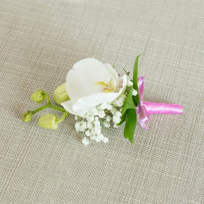 A carefully crafted orchid boutonniere lies horizontally across an oatmeal-tone linen backdrop, its placement and soft, even lighting giving the impression of a quiet styling moment before it is pinned to a lapel for a Stanmore celebration. The fabric beneath has a subtle woven texture, warm and neutral, allowing every detail of the flowers to stand out clearly. At the very centre of the piece, a single large white Phalaenopsis orchid opens with serene confidence, its wide petals velvety and unblemished, curving gently outward like calm, unfurled wings. The throat of the orchid glows with a delicate wash of yellow and a hint of pale pink, adding depth and warmth to the otherwise cool white bloom. Nestled close to its base, cloud-like clusters of white baby's breath create a cushion of tiny, rounded flowers, softening the lines of the orchid and lending a light, romantic texture that feels timeless. Just behind, a few slender leaves in vivid green slip into view, their clean edges and natural gloss giving the arrangement a fresh, garden-picked feel, as though gathered that morning from a sheltered corner of a Stanmore back garden or near Bentley Priory Nature Reserve. To one side, a short side stem reaches out, bearing two small, tightly closed orchid buds in pale lime-green, poised and smooth, echoing the promise of future blossoms and lasting memories. The stems are cinched together at the base with a bright fuchsia pink satin ribbon, tied into a neat bow whose lustrous surface gently reflects the light, providing a joyful contrast to the neutral background. It is the kind of elegant, handcrafted boutonniere suited to a groom, groomsman, or prom guest, perfect for formal occasions across Stanmore, from intimate home gatherings to receptions along The Broadway.