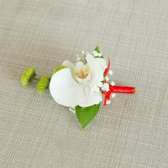 An elegant boutonniere, carefully designed for a wedding or black-tie event, is shown resting on a softly textured, light beige linen surface that evokes the understated charm of a well-set table at a Stanmore reception. At its centre, a single white phalaenopsis orchid blossom unfolds with serene confidence, each broad petal smooth and luminous, creating a pristine frame around the delicately patterned throat. The inner heart of the orchid glows with gentle yellow tones that fade into fine streaks of reddish-orange or warm brown, adding depth and a sense of quiet drama without overshadowing the bloom's pure white form. Around the orchid, sprays of airy white baby's breath are tucked in like little clouds, their tiny flowers softening the lines of the design and introducing a light, romantic texture reminiscent of morning mist over Bentley Priory Nature Reserve. To the left, two spherical lime-green chrysanthemum-style buds stand out with their dense, spiky texture, injecting a bright, modern accent that contrasts beautifully with the orchid's classic elegance. A rich green leaf fans out beneath the arrangement, anchoring the design and giving it a verdant base, while a smaller, darker green leaf peeks from near the stem, enhancing the layered, natural feel. The stems themselves are bound in a vivid crimson red satin ribbon, wrapped snugly and finished in a neat little bow or knot, catching the light with a subtle sheen and adding a romantic splash of colour. The overall effect is sophisticated yet fresh, capturing the feeling of a thoughtfully chosen buttonhole ready to be pinned to a lapel for a Stanmore wedding, prom, or formal celebration where every small detail matters.