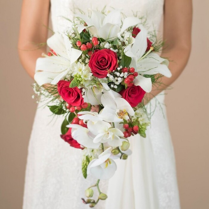 A Stanmore wedding is beautifully expressed in this richly detailed image: a bride, partially visible in her white lace gown, cradles a generous teardrop bouquet that flows from her hands in a vibrant display of craftsmanship. The bouquet's stars are the bold white oriental lilies, their broad, open petals and distinctive yellow stamens reaching out in stately elegance. A stream of white phalaenopsis orchids cascades from the centre, each bloom showcasing intricate centers and smooth, trailing stems, imparting a feeling of movement and luxury. Scattered among these, crimson red roses add drama and depth, their plush petals unfolding with promise. Clusters of glossy red hypericum berries and delicate wisps of baby's breath and tiny white filler blossoms lighten the mood, providing both structural variety and a playful texture. A framework of finely-textured asparagus fern and deeper, supportive green leaves firmly root the bouquet in the area's green, leafy character. The image's background is purposefully understated-a gentle, blurred beige-ensuring the bouquet's colours and intricate structure remain the visual highlight. Local Stanmore florists have infused this arrangement with thoughtful artistry and vibrant energy, making it an inspired choice for weddings or receptions taking place in Stanmore's beloved historic venues or tranquil gardens.
