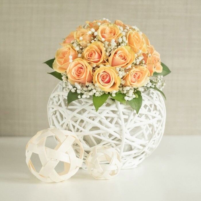 A sophisticated floral composition is arranged as a low, rounded centerpiece, featuring a generous cluster of soft peach and apricot roses, tightly grouped so their petals almost touch, forming a smooth, domed silhouette. Each rose reveals a subtle gradient of colour, with richer, warm orange tones nestled in the centre and lighter, cream-infused peach at the edges, like the glow of late afternoon light over the quiet streets of Stanmore. Between the rose heads, delicate sprays of white baby's breath (gypsophila) fan out lightly, their tiny blooms catching the light and softening the overall look, while hints of deep green foliage peek from beneath, lending structure and a natural frame. The arrangement is cradled in a striking white spherical vase, meticulously crafted from interlaced strips or strands that create an open, airy lattice pattern, allowing glimpses into the interior and adding a strong textural contrast to the smooth rose petals. Resting on the immaculate white tabletop in front of the vase are two small, decorative woven spheres in a similar pale white tone, one slightly larger and more geometric, the other looser and more abstract, echoing the artistic feel of the main container. Behind, a lightly textured wall in soft beige or greige forms a serene backdrop, free of clutter, so the colours and craftsmanship remain the focus. The even lighting suggests gentle daylight, as if this centerpiece were placed near a bright window in a Stanmore living room or a boutique venue off The Broadway. Created with the finesse you'd expect from a local expert like Flower Delivery Stanmore, the scene evokes refined celebration, perfect for weddings, intimate dinner parties, or elegant home décor across the Stanmore area.