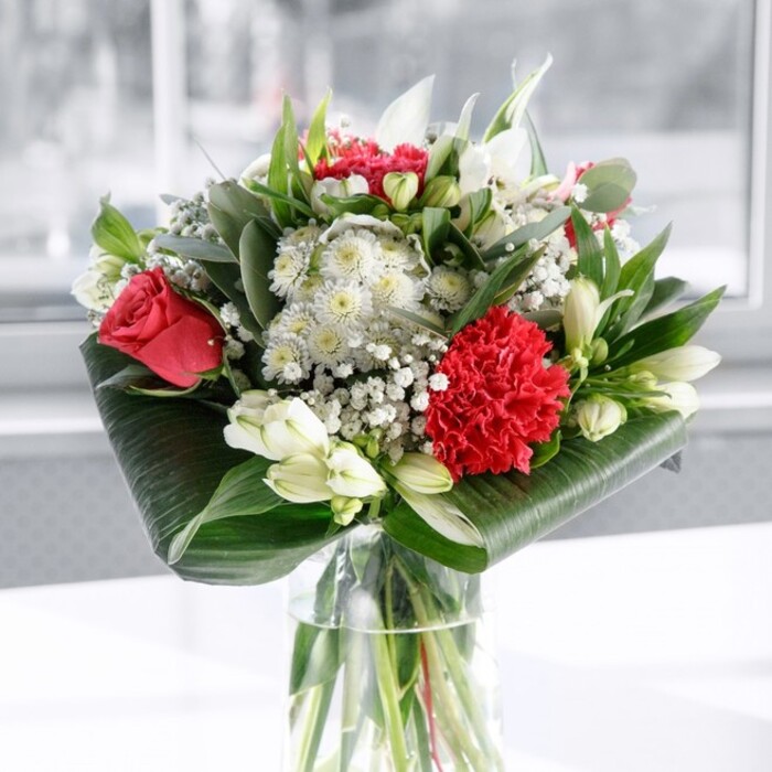 In this image, a carefully crafted bouquet of red and white flowers draws the eye immediately, standing tall in a simple, clear cylindrical glass vase. Through the vase, you can see straight, freshly cut green stems fanning out in the water, suggesting a bouquet that has just been arranged with precision and care. The main drama comes from deep red roses and a prominent crimson carnation, their velvety petals and ruffled edges creating a rich, romantic focal point. The carnation, with its many soft, frilled layers, sits near the front, adding distinctive texture, while the roses rise slightly above, each bloom opening enough to show off its classic spiral form without yet being fully blown. Interwoven between these reds are pure white and creamy blossoms: alstroemeria with long, narrow petals marked by delicate interior speckles, snowy daisy-like flowers, and several white lily buds still closed, their smooth, tapered forms suggesting elegance and potential. Fine sprays of baby's breath float gently throughout, their tiny white blooms bringing an almost cloud-like softness and helping to fill the spaces without crowding the arrangement. Encircling this mix of blooms is a generous frame of greenery. Broad, glossy leaves in a deep, almost lacquered green are wrapped and curled neatly around the base, forming a structured collar that feels both modern and classic. Above them, slimmer green stems and eucalyptus-style foliage rise to add contrast in shape and shade. The bouquet rests on a clean white surface, the background softly blurred and bright, evoking natural daylight spilling into a Stanmore home near Whitchurch Lane or overlooking Bentley Priory Nature Reserve. The overall impression is of thoughtful refinement, a bouquet that could easily grace a dining table for an anniversary dinner, a Mother's Day brunch, or a special celebration with friends, bringing a sense of warmth and polished beauty into the room.