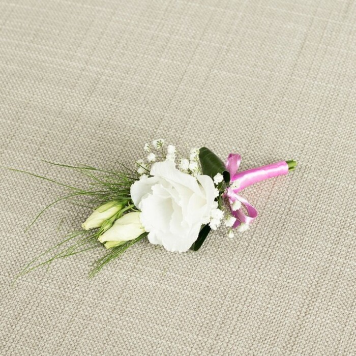 A small, elegant boutonniere lies at a diagonal angle on a warm, light beige fabric that resembles tightly woven linen or soft burlap, its subtle weave giving the scene a natural, tactile charm. The central focus is a single white lisianthus in full bloom, its ruffled petals layered in soft, rounded tiers that open out like a delicate rosette. The white is clean and bright, yet softened by faint creamy tones towards the centre, as if the flower has just opened that morning for a wedding somewhere in Stanmore. Close beside it, on the lower left, are two slender lisianthus buds, still closed and tinged with pale green, their smooth, tapered shapes echoing the main flower while suggesting life still unfolding. Interspersed among the bloom and buds are tiny clusters of white gypsophila, the baby's breath forming small, airy puffs that lend the arrangement a classic bridal feel. From behind the flowers, fine, grass-like blades of dark green foliage splay gently outwards towards the left, adding line and movement, while a single broader, deeper green leaf cups the back of the lisianthus, providing depth and contrast. The stems are neatly aligned and gathered at the lower right, wrapped in a bright fuchsia pink satin ribbon that has been tied into a petite, neatly formed bow. The vivid pink stands out joyfully against the muted background, its sheen catching the soft, even light that falls across the scene. The overall impression is of a fresh, handcrafted boutonniere, perfect for a groom, best man, or prom guest arriving from nearby Canons Park or along The Broadway in Stanmore, bringing a quiet touch of garden elegance to a formal outfit.