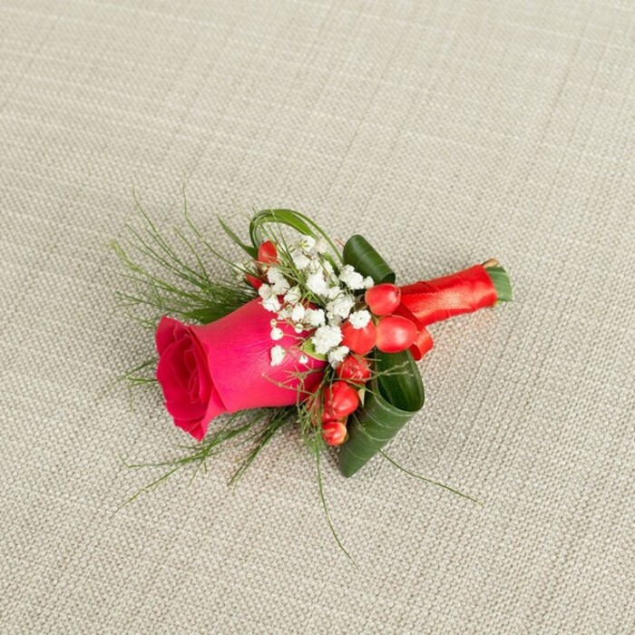 A small, elegant floral boutonniere rests at a gentle diagonal on a pale beige, lightly textured fabric backdrop, evoking the calm of a dressing room in Stanmore just before a ceremony begins. The arrangement's focal point is a single, striking deep fuchsia rose, its tightly layered petals forming a lush spiral that gradually relaxes towards the outer edges. The rose appears freshly cut, with a velvety, almost luminous quality to its colour, as if just prepared for same-day wear. Around the base of this rose, airy clusters of white baby's breath gather in delicate sprays, their tiny blossoms sprinkled like confetti, softening the strong pink and lending an almost bridal touch. Bright red hypericum berries are woven among these white flowers, each berry plump and polished, providing a festive pop of colour that suggests warmth, celebration, and the joyful atmosphere of a wedding reception at a local Stanmore venue. The greenery is thoughtfully varied: fine, whispery fronds of fern-like foliage extend outward, adding movement and a sense of natural looseness, while broader, darker green leaves are carefully curled and layered to create a supportive cradle around the rose, giving the piece structure and depth. The stems are gathered together and expertly bound in a smooth, satin red ribbon, wrapped firmly and tied into a compact, graceful bow that echoes the rose's rich hue. Soft, even lighting falls from above, revealing subtle shadows that accentuate each petal edge, leaf curve, and berry highlight. The overall effect is of a refined, handcrafted floral detail-perfect for a groom, groomsman, or formal event guest walking through Stanmore's tree-lined streets on a day filled with love and promise.