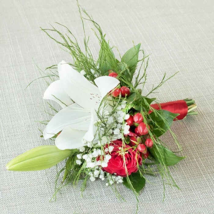 Crafted by expert florists in Stanmore, this sophisticated small arrangement, perfectly suited for a wedding or anniversary, radiates both classic beauty and local warmth as it rests on a subtly woven light beige fabric. The focal point-a majestic white lily-opens widely to reveal delicate reddish-brown speckles and sunny yellow anthers, evoking a sense of purity and natural elegance. Adjacent, a rich, velvety red rose exudes passion and depth, its petals tightly curled in a timeless expression of love. Threads of white baby's breath meander throughout, softening the composition with a fine, airy touch, while vibrant clusters of glossy red berries sparkle against the creamy tones of the lily. These highlights bring to mind festive occasions celebrated throughout Stanmore's close-knit community. A slender green lily bud arcs gently to the side, a quiet symbol of promise and renewal. Surrounding the central blooms, a medley of textured foliage-from feathery fern fronds to bold, polished leaves-provides fullness and a miniature landscape reminiscent of Stanmore's verdant green spaces. The expertly tied red satin ribbon at the stems adds a finishing flourish, its sheen catching the soft light. Under even lighting, the bouquet's fresh colours and graceful details are showcased, making it a stunning choice for local weddings, anniversaries, or as a refined gift delivered by Flower Delivery Stanmore.