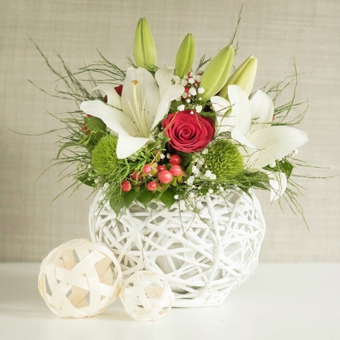 This image shows an expertly crafted floral centerpiece designed for a wedding or formal dinner in Stanmore, illuminated by soft, even light that emphasizes every petal and texture. At the base of the arrangement is a round white vase with an intricate, open-weave construction, its overlapping strands forming a lattice that recalls contemporary basketry. Rising from this vessel is a lush composition dominated by white lilies and red roses, accented by fresh greenery and unusual green pom-pom blooms. Several lilies have opened wide, their petals smooth and slightly curved at the edges, creating trumpet-like forms that draw the eye inward to pale freckles and a cluster of golden stamens. Other lilies remain in slender, closed bud form, suggesting continued blooming long after the event. Close to the centre, a particularly striking red rose is showcased, its velvety petals arranged in a tight spiral rosette that symbolises enduring affection and stands out boldly against the white lilies. Surrounding these main flowers, bright green spherical blooms-likely green dianthus or button chrysanthemums-create little cushions of texture, their vivid colour reminiscent of fresh grass in Canons Park on a clear day. Sprays of tiny white gypsophila weave gently through the larger flowers, providing a light, cloud-like softness. Small clusters of dark red or reddish-brown berries punctuate the greenery, adding seasonal warmth and a touch of rustic charm. A variety of foliage spills over the edge of the vase, from fine, feathery fronds that arc gracefully outward to broader, darker leaves that ground the design and frame the white and red blooms. The vase rests on a clean white surface that subtly reflects light, enhancing the arrangement's brightness. To the left, two decorative woven balls in a pale, natural wood colour sit slightly in front of the vase, echoing its texture and reinforcing the cohesive, contemporary yet organic styling-a perfect fit for wedding tables and celebratory gatherings across Stanmore.