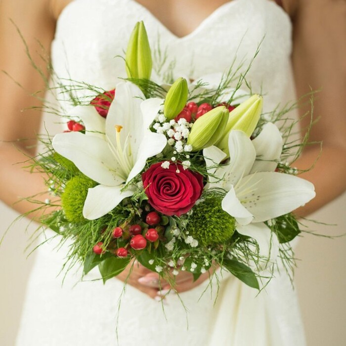 A bride's moment in Stanmore is beautifully encapsulated in this close-up, where elegance and vibrant colour come together. Her white strapless gown, featuring a subtle, patterned fabric, flows with understated grace behind the bouquet she holds. Dominating the arrangement are snow-white oriental lilies-some fully open to reveal striking golden stamens, others tightly closed as fresh green buds, representing both present celebration and future promise. Nestled at the core, a deep red rose clenches its tightly furled petals, a true icon of romance and enduring love that resonates throughout Stanmore's wedding traditions. The bouquet is further enriched by delicate clusters of baby's breath, bright green clusters of trick dianthus or similar blooms, and brilliant red hypericum berries, each providing visual interest and texture. Framing these highlights is an abundance of lush greenery, from slender asparagus fern fronds to broader, glossy leaves, giving the bouquet a rounded, natural fullness akin to Stanmore's well-tended parks and gardens. Under soft light, every nuance of petal, leaf, and pattern is revealed, creating a vivid impression of local artistry and bespoke floral design accessible to brides across Stanmore and its surrounding areas.
