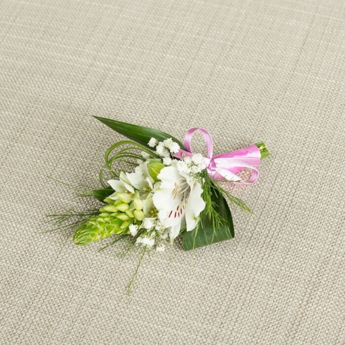 An elegant boutonniere, carefully arranged as though ready to be pinned to a groom's lapel in Stanmore, rests against a subtly textured, light beige fabric that resembles fine natural linen. At the centre of the composition is a single white alstroemeria, sometimes called a Peruvian lily, its petals broad, slightly ruffled, and delicately veined. Near the throat of the flower, fine reddish-brown streaks and speckles radiate outwards, offering intricate detail that contrasts tastefully with the pure white of the petals. To the left side, a vibrant lime-green bud cluster rises like a small, tapered spike, each tightly packed bud lending a sculptural presence and a sense of youthful freshness, as if moments from opening. Threaded around and between these key elements are airy sprigs of white baby's breath, their tiny blossoms forming a soft halo that lightens the overall design. The florist has balanced this delicacy with a foundation of rich greenery: broad, dark green, pointed leaves support the flowers from beneath, while long, slender blades of grass-like foliage curve gently outward, giving the boutonniere a natural, flowing silhouette. Securing the stems on the right is a neat bow of glossy pink satin ribbon, its smooth texture and cheerful colour adding a subtle note of romance and festivity. The lighting is even and gentle, with no harsh shadows, allowing the viewer to appreciate the interplay of textures-from the crisp flower petals to the woven fabric beneath. The arrangement feels timeless yet contemporary, perfectly suited for weddings, proms, or anniversary dinners across Stanmore, whether worn at a ceremony near Canons Park or presented as a thoughtful finishing touch arranged by a trusted local florist.