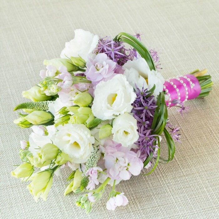 Laid gracefully on a soft, neutral-toned linen, this hand-arranged bouquet is a vision of refined celebration, blending the natural elegance of Stanmore with artistic floral craft. At the centre, lush and creamy white Lisianthus open in plush ruffles, each petal surrounding a gentle yellow centre, their beauty heightened by elegant elongated buds in white and lime green. These pure tones are perfectly complemented with pastel lavender-pink stock flowers, which create a soothing cloud-like texture, and by vibrant deep purple allium, their striking, spiky star forms evoking a playful sparkle. Splashes of green-slender bear grass curves and a profusion of lime-tinted buds-infuse the bouquet with depth and freshness, reminiscent of the rolling greenery at Stanmore's edges or a stroll through Bernays Gardens. The bouquet's stems are wrapped neatly with hot fuchsia satin ribbon and encircled by a strand of polished white pearls, merging classic style with modern vibrancy. Designed to shine at spring weddings or elegant gatherings, this bouquet channels the local spirit-romance, unity, and timeless beauty-right from Stanmore to special moments celebrated everywhere. It's more than flowers; it's an experience, echoing the subtle colours and tranquil charm of its local origins.