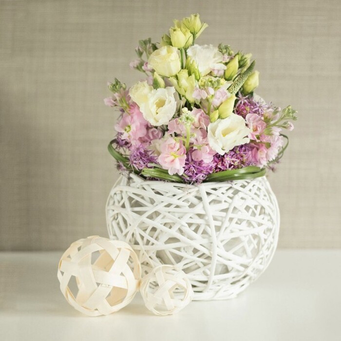 In Stanmore, where refined taste meets natural beauty, this wedding table arrangement unfolds as a symphony of pastel florals and creative texture. The bouquet features layers of creamy white Eustoma, their elegantly ruffled petals touched with hints of pale yellow that glow beneath soft daylight, all complemented by upright clusters of soft pink and lavender stocks for a vertical flourish. At the base, rich purple filler flowers-perhaps local asters-provide a tactile, colour-rich foundation, bordered by lush, cool-green foliage akin to the landscapes surrounding Bernays Gardens. The centerpiece is cradled in a contemporary, spherical vase, its open lattice of painted white rattan or delicately woven branches echoing the hand-crafted sensibility that characterises Stanmore's neighbourhood florists. Two matching decorative spheres-created from pale, airy rattan-rest to the side on the smooth, white surface, their geometric weaving amplifying the arrangement's modern artistry. Framed by the gentle texture of a warm beige wall covering, the setting is bathed in tranquil natural light, resulting in an atmosphere of fresh luxury and understated elegance. Commissioned by Flower Delivery Stanmore, this arrangement offers not only visual beauty but also a spirit of celebration and community, perfect for wedding tables or sophisticated events in Stanmore.