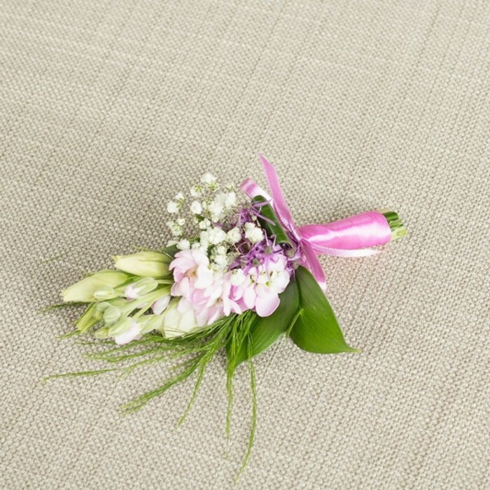A refined floral boutonniere or petite corsage is presented against a softly woven linen surface in a warm, oatmeal-beige tone, the fabric's gentle texture evoking a table runner or suit fabric chosen for a Stanmore wedding. The neutral background allows every nuance of the flowers to stand out, from the delicate blush of the petals to the rich green of the foliage. At the heart of the arrangement, several ruffled soft pink blooms-reminiscent of miniature stocks or small cottage-garden flowers-are clustered together, their petals tightly layered and slightly tousled, creating a sense of fullness despite the small scale. Interlaced with these pink flowers are fine sprays of classic white baby's breath, the tiny star-like blossoms scattered along thin stems to form a light, cloud-like halo around the central cluster. Adding depth, small accents of lavender or soft purple florets are tucked discreetly among the pinks and whites, giving the eye a subtle pop of cooler colour without overpowering the design. To one side, a graceful row of creamy white to pale green buds, perhaps freesias or similarly slender blooms yet to open, seems to stretch outwards in a quiet arc, implying movement and the promise of future blossoms. The florals are cradled by broad, glossy green leaves that create a sturdy base, while wispy, grass-fine fronds and fern-like sprigs extend outwards, lending an organic, slightly whimsical feel, like greenery gathered from gardens around Bernays Gardens or the edges of Stanmore Common. At the base, the stems are neatly gathered and bound in a vivid fuchsia satin ribbon, tied into a precise, symmetrical bow with softly curved loops and neat tails resting against the linen. The gentle, even lighting highlights the satin's sheen and the velvety quality of the petals, capturing a sense of fresh preparation, as if this piece has just been finished in the Flower Delivery Stanmore workshop and set down before heading out for same-day delivery to a local wedding, prom, or elegant family celebration.