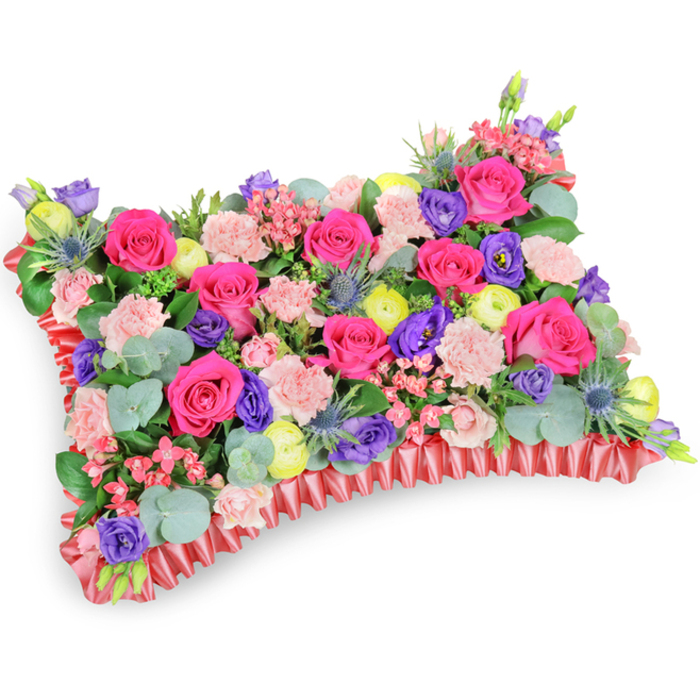 Presented against a stark white background from a slightly oblique, elevated angle, this pillow-shaped floral tribute is densely packed with fresh blooms and varied foliage, each element chosen for colour, texture and sentiment. The principal notes are hot pink roses, their velvety petals forming the dominant, jewel-like presence across the top surface. Interspersed are pale pink carnations, their frilled petals adding a soft, tactile contrast, while deep purple lisianthus provide sculptural depth and a graceful silhouette. Lemon-yellow ranunculus and small creamy accents lift the palette with bright, hopeful points of colour, and the inclusion of silver-blue eryngium thistles introduces a striking, spiky counterpoint that adds architectural interest. Eucalyptus leaves, rounded and silvery-green, weave through the composition alongside darker green fillers, supplying structure and a cool, resinous scent that tempers the sweeter floral notes. The entire cushion is bordered with a rich red satin ribbon, worked into a pleated, ruffled edge that clearly defines the rectangular shape and lends a formal, commemorative finish. Under soft studio lighting the arrangement shows subtle highlights on petal edges and ribbon folds, creating gentle shadows that enhance the three-dimensional quality. This is the kind of sympathy pillow often chosen for funeral tributes or graveside memorials in the Stanmore and Harrow area - fitting for placement near Stanmore tube or at a service close to Stanmore Park - where the vibrant yet comforting combination of colours conveys remembrance, dignity and a celebration of life.