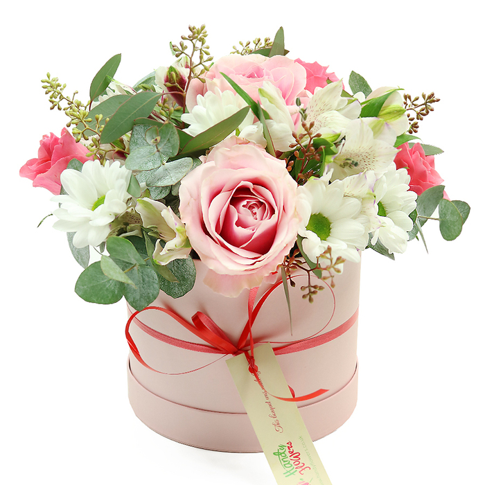 A chic, round hatbox in soft pastel pink supports a rich, domed bouquet of mixed flowers, photographed against a bright white backdrop that gives the composition a fresh, airy feel reminiscent of light-filled homes around Stanmore. The cylindrical box has a smooth, matte surface and is accented with a narrow coral-red satin ribbon, neatly tied into a small bow on the front so the ribbon's sheen catches the light. From the bow, a cream rectangular gift tag hangs delicately, its surface printed with refined green script and a website address, subtly reinforcing the idea of a bespoke florist delivery. Above the rim of the hatbox, flowers appear to burst gently outward, arranged with an eye for both balance and softness. A large, fully opened rose sits prominently near the front, its petals shading from very pale blush on the outer edges to a deeper pink spiralling elegantly toward the centre, giving the impression of depth and movement. Surrounding it are white, daisy-like chrysanthemums with bright yellow-green centres, lending structure and a touch of innocence. Nestled among them, white alstroemeria blooms show off their trumpet shapes and fine inner speckles, while clusters of smaller, vibrant deep-pink flowers-possibly spray roses or lisianthus-add pops of saturated colour that draw the eye across the arrangement. Eucalyptus leaves in muted silver-green tones arc between the blooms, mingling with darker green foliage and slender twigs tipped with tiny green buds and muted brown elements, creating layers of texture. The overall look is refined yet inviting, suited to marking special occasions like birthdays, anniversaries, or Mother's Day gatherings in Stanmore and nearby Edgware, and conveying a quiet message of love, appreciation, and thoughtful care.