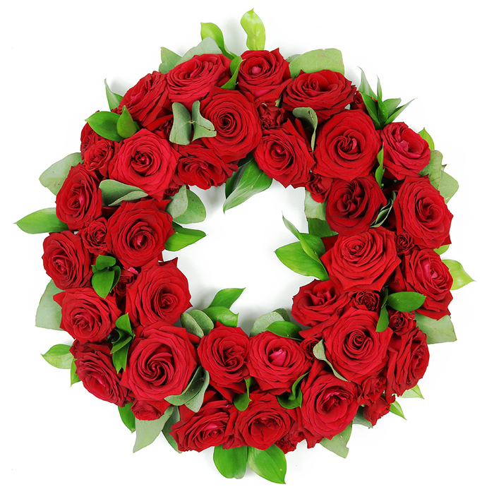 An artful circular wreath of deep red roses rests against a crisp white backdrop, a testament to studio craft by a Stanmore florist. The wreath is densely packed with dozens of crimson blooms in various stages of opening, from tightly furled buds to elegantly unfurled, velvety petals, each displaying rich, saturated hues and intricate folds. These abundant red roses, traditionally symbolizing love, remembrance, and respect, create a continuous, unbroken ring. Interspersed throughout the lush bloom are clusters of fresh green foliage: broad, matte eucalyptus-like leaves provide a soft, verdant frame, while slender, glossy ruscus leaves add sharp, emerald accents that elevate the texture and depth. The careful arrangement gives the wreath a full, three-dimensional quality, with layers of petals and leaves subtly overlapping. This elegant red rose wreath conveys a profound message of solemn beauty and heartfelt tribute, a powerful visual element often seen at memorial services, funeral arrangements, and commemorations. The striking combination of passionate red and serene green, set against a pure white backdrop, makes this floral wreath a poignant representation of remembrance and respect, suitable for delivery across Stanmore's neighbourhoods, Canons Park, and surrounding streets.