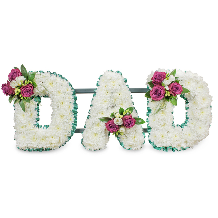 This elegant sympathy piece spells out the letters DAD in capital form, each letter lovingly constructed from a thick layer of pristine white chrysanthemums that give a velvety, tactile surface against a stark white background. The tiny, cushiony florets nestle together to form bold, readable shapes whose soft petals diffuse light and suggest a sense of peaceful clarity. A vibrant emerald-green ribbon-pleated or gently ruffled-traces the perimeters and inner openings of each letter, creating a clear, solemn outline and a refined visual contrast to the white blooms. A discreet dark green or grey bar runs behind the letters, implying the easel or support that will carry this design to a funeral, memorial service or graveside.

Thoughtful accent clusters of deep pink to magenta roses, paired with delicate white freesia and full, glossy foliage, are positioned to balance the composition: the first D bears its cluster at the upper left curve, the A is graced with a group in its lower V, and the final D reflects the first with a cluster on its upper right curve. The roses lend a plush, tactile warmth while freesias add a soft citrus-honey scent; the greenery brings contrast and structure. I can imagine this piece positioned in the quiet light of a chapel near Stanmore Broadway or set beside a headstone close to Stanmore tube station, offering a clear, heartfelt expression of love, respect and farewell.
