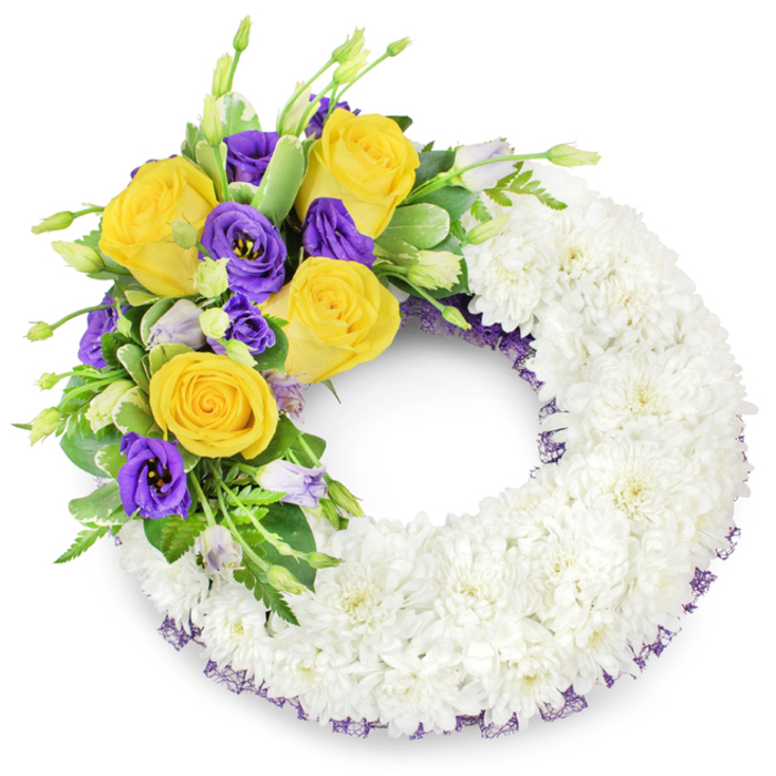 An elegant, circular sympathy wreath set against a plain white background, the kind of piece our team at Flower Delivery Stanmore would assemble for a respectful funeral tribute near Stanmore High Street or Bentley Priory. The principal structure is a dense, continuous ring of small white spray chrysanthemums, closely packed to create a plush, cloud-like texture that suggests purity and collective remembrance. On the upper left quadrant an accent cluster curves gracefully over the white base: warm yellow roses, their petals unfurling in layers of butter and gold, are arranged with considered spacing so each bloom has presence. Contrasting these are deep purple lisianthus with softly ruffled, cup-shaped blossoms that lend a velvety coolness; scattered unopened buds in pale lavender and green introduce a sense of life and progression. Mixed greenery - glossy broad leaves, slender fern fronds and touches of variegated foliage - frames the colours and adds natural movement, helping the cluster sit gently atop the wreath's surface. A narrow dark purple mesh trim peeks from beneath the flowers along the inner and outer edges, a subtle detail that ties the composition together and echoes the lisianthus tones. Under soft, neutral lighting the textures and hues read clearly: the chrysanthemums' soft mass, the roses' warm radiance, and the lisianthus' dignified depth. The design communicates condolence, quiet beauty and a composed farewell suited to memorial services across Stanmore and nearby neighbourhoods.
