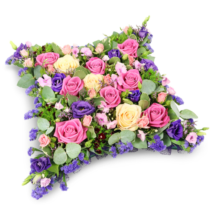 A meticulously arranged floral cushion tribute fills the frame, its pillow-like form created from a tightly packed mixture of roses, lisianthus, filler flowers, and lush foliage. The cushion is rectangular with gently rounded corners, slightly raised in the centre so it appears soft and inviting, symbolic of rest and repose. Across the surface, roses are the star: bold fuchsia blooms sit beside softer pastel pink roses, their many petals opening into rounded, full heads. Ivory-cream and pale yellow roses are interspersed among them, catching the light and bringing a gentle warmth to the palette, like a soft glow in a quiet room. Between the roses, deeper violet and royal-purple lisianthus provide contrast, their ruffled petals and more elongated heads adding movement and texture. Smaller blush spray roses and tiny lavender-blue filler flowers, probably statice, ensure that there are no gaps, giving the cushion a luxuriantly full, almost quilted appearance. A base of greenery cradles the flowers: rounded, silvery eucalyptus leaves introduce a cool, misty tone, while more delicate, fern-like foliage in richer green peeks out here and there, softening the outline and adding dimension. The tribute is photographed against a plain white background with a subtle shadow beneath, so its refined craftsmanship and colour harmony stand out clearly. This type of floral pillow would be a fitting tribute for a funeral or memorial service in Stanmore, perhaps at a crematorium near Bentley Priory or a chapel along The Broadway, chosen by relatives who want to send a message of comfort, peace, and enduring love. It carries the quiet dignity and heartfelt sentiment that many families seek when saying a final, respectful goodbye.