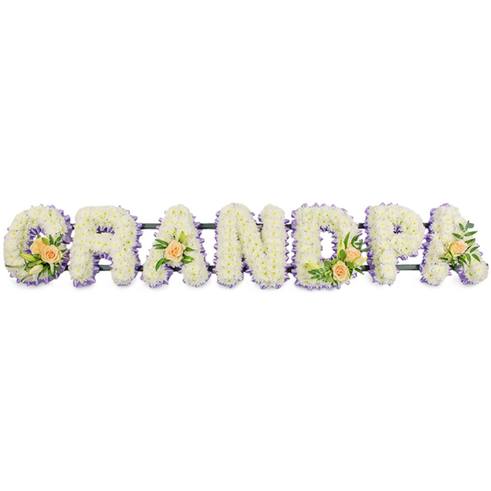 In this detailed floral name tribute, the word GRANDPA is spelled out in large, upright letters that sit in a straight horizontal line against a crisp white background, reminiscent of a tribute prepared for a quiet Stanmore chapel or cemetery service. Each capital letter is meticulously filled with small white chrysanthemum heads or similar blooms, placed so closely together that the surface appears thick and luxurious, with subtle shadows between the petals adding depth. Around the perimeter of each letter runs a delicate edging of soft lilac or pale purple flowers, which gently outlines the forms of the G, R, A, N, D, P, and A, making them easy to distinguish from one another and from the light backdrop. The tribute is further personalised with the inclusion of peach roses, carefully positioned so that they add warmth without overpowering the composition. On the leftmost letter G, a cluster of peach roses, some fully open and others still in tight bud, is accompanied by slender white lily-style buds and glossy green foliage, creating a focal accent. The first A holds a single peach rose nestled in greenery, while the central D echoes the G with a second cluster of roses and leaves. The final A repeats the single-rose motif, bringing visual harmony to the right side of the tribute. All letters are fixed onto a narrow, dark horizontal bar, probably fabricated from metal with floral foam, which provides discreet support and keeps the arrangement aligned for display on stands at a funeral home, church, or graveside in and around Stanmore, from Belmont Circle to the quiet roads near Stanmore Country Park. The overall palette of white, lilac, and soft peach communicates a message of peace, affection, and heartfelt thanks to a cherished grandfather.