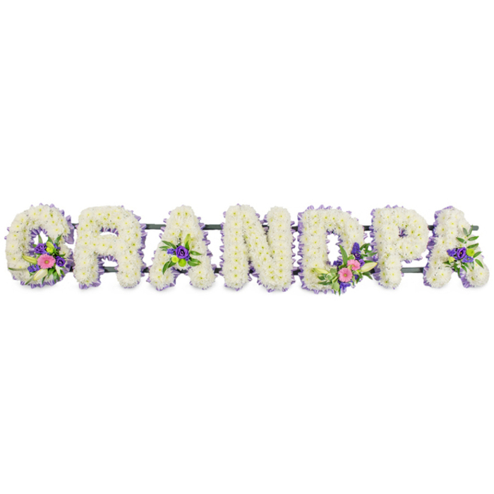 In this photograph, the word GRANDPA is lovingly spelled out in large floral letters, presented in a straight line against a clean, white studio backdrop. Each capital letter is expertly constructed from dense clusters of small white chrysanthemums, their layered petals forming a plush, almost quilted surface that appears soft to the touch and symbolises peace and remembrance. Running neatly around the perimeter of each letter is a slim border of lilac and pale purple blossoms, most likely tiny statice or similar filler flowers, giving definition to the shapes and creating a gentle contrast without overwhelming the calm white core. Scattered through certain letters are artfully arranged accent groupings: the G hosts a cluster of elegant white lily-style flowers surrounded by bright pink and fuchsia blooms and deeper purple highlights; the first A and the final A hold smaller, equally thoughtful clusters; and the D contains one of the more prominent focal sprays, complete with vibrant pinks, whites, purples, and touches of fresh green button chrysanthemums and foliage. A slim, dark green frame runs quietly behind the line of letters, indicating the supportive stand that would allow this tribute to be displayed upright at a funeral in Stanmore, perhaps at a church on Marsh Lane or a nearby crematorium. The lighting is soft and even, helping the viewer notice every ruffled petal and subtle change in colour, while the white background removes any distraction from the heartfelt message spelled out in flowers. The design exudes dignity and tenderness, a bespoke sympathy arrangement that would serve as a meaningful centrepiece at a memorial service, honouring a beloved grandfather with elegance and care.