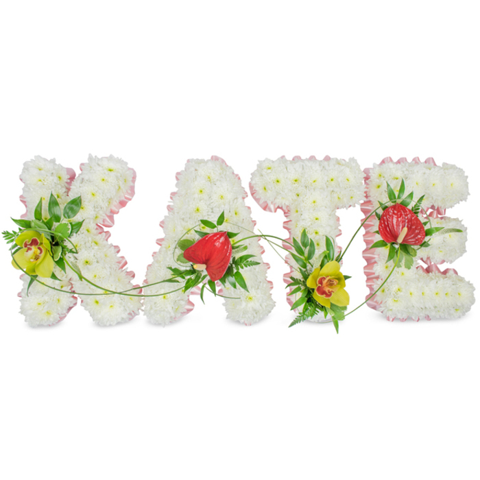 In this photograph, the name KATE appears as a refined floral composition, each letter carefully formed from fresh white chrysanthemums and presented against a crisp, pale background. The flowers are densely arranged so that no gaps show, creating large, block-style letters with a plush, cushiony feel. A narrow band of soft pink satin ribbon traces the perimeter of every letter, gently rippling as it catches the light and offering a sweet, understated contrast to the white blooms. At the upper left of the K, a yellow-green cymbidium orchid with a speckled red-brown throat is set among glossy dark leaves and delicate fern fronds, while a matching orchid nestles at the base of the T, providing visual balance and rhythm across the tribute. The A and E each carry a single, heart-shaped red anthurium bloom, their high-gloss, waxy surfaces and bold red colour standing out as symbols of love and strength. Slender green stems and curling tendrils drift between the letters, lightly resting over the chrysanthemums so that the name feels linked and flowing rather than rigid and separate. The even lighting and uncluttered background draw attention solely to the craftsmanship and harmony of the design. It is easy to imagine this KATE arrangement placed at the front of a Stanmore funeral service or by a graveside at Harrow Weald, chosen by family members who wanted something personal yet tasteful. The piece conveys respect, affection, and gentle comfort through its colour palette, textures, and careful placement of each accent flower.
