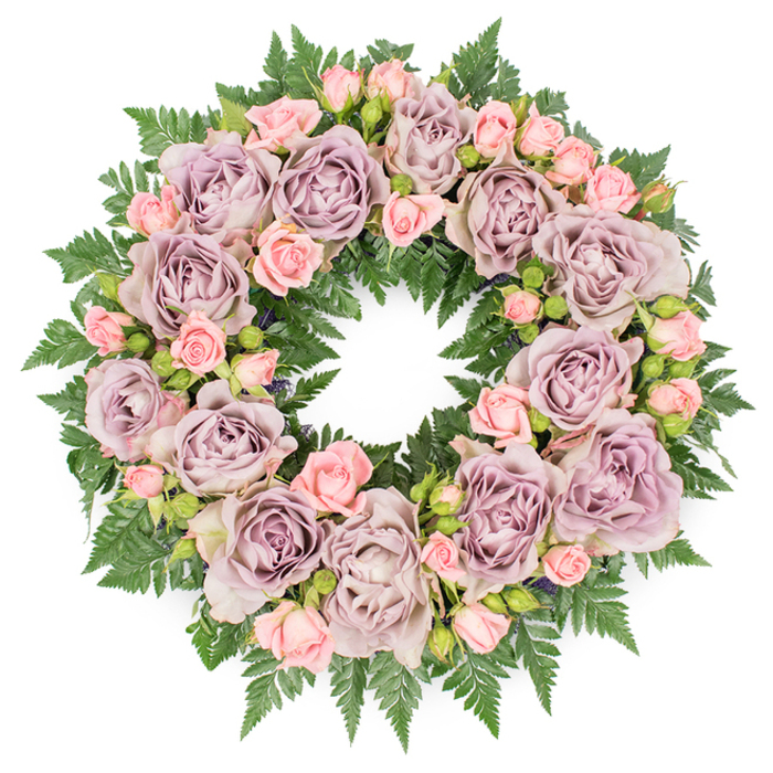 An elegant sympathy wreath rests flat on a stark white background, its flawless circular silhouette immediately conveying wholeness and continuity. The composition is lush yet controlled, crafted with the sensitivity of a florist who understands the quiet language of farewell flowers. Large, fully open roses in tones of dusty lavender and muted mauve create the primary visual rhythm, placed evenly around the ring so that no section feels heavier than another. Each of these roses reveals layers upon layers of soft petals, the outer edges slightly loosened, suggesting a calm, graceful maturity. Interspersed among them are clusters of smaller blush pink spray roses, some still in firm bud, others in gentle half-bloom, adding a lighter note that softens the deeper purple hues. This interplay between the larger, stately roses and the delicate, youthful sprays introduces both visual texture and emotional nuance, like different voices in a shared remembrance. Beneath this tapestry of blooms lies a dense collar of dark green ferns, most likely leatherleaf or a similar variety, their feathery, serrated leaves overlapping to create a rich, verdant foundation. The foliage extends just beyond the flowers, forming a natural, leafy halo that frames the entire wreath and contrasts beautifully with the clean white background. The overall lighting is even and subdued, bringing out the soft lustre of the petals and the matte texture of the leaves without harsh glare. This wreath would feel entirely at home delivered to a Stanmore funeral service or laid gently at a graveside, its gentle colours and thoughtful design offering a message of peace, gratitude, and dignified farewell for a loved one remembered with affection.