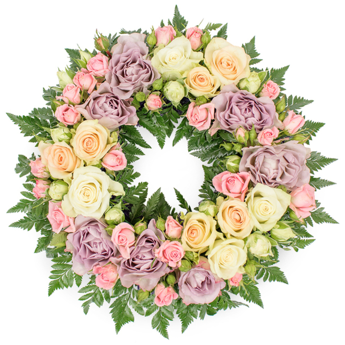 A delicately balanced circular wreath of roses and fern greenery is placed against a smooth, white background, the neutral setting allowing every flower and leaf to be fully appreciated. The wreath's surface is densely covered with roses, so the eye moves from bloom to bloom without interruption, tracing a complete ring of soft colour. Ivory and creamy white roses provide a gentle base tone, interwoven with pale peach and buttery yellow blooms that lend warmth, while blush pink roses and distinctive dusty lavender or muted mauve flowers introduce a romantic, vintage nuance. Many of the roses are fully open, their layered petals unfurling in wide, cupped shapes, while others remain in half-open or tight bud stages, giving the design texture and a natural, evolving feel. Scattered among them are smaller pink spray roses and dainty, pale buds that fill the spaces and soften every transition. Beneath the petals, lush, dark green fern fronds create a supportive framework, their finely cut leaves appearing between flowers and around the outer edge, adding depth and a cool, woodland contrast to the otherwise pastel palette. The wreath's perfect circular form suggests eternal love, ongoing remembrance, and the continuous nature of family ties, themes that resonate strongly at funerals and memorials across Stanmore, from quiet residential streets to services held near Canons Park. The overall mood of the arrangement is serene and contemplative, yet undeniably rich, a fitting choice for loved ones wishing to express their sympathy with a floral tribute that feels both traditional and gently contemporary, crafted with the careful attention of a local Stanmore florist.
