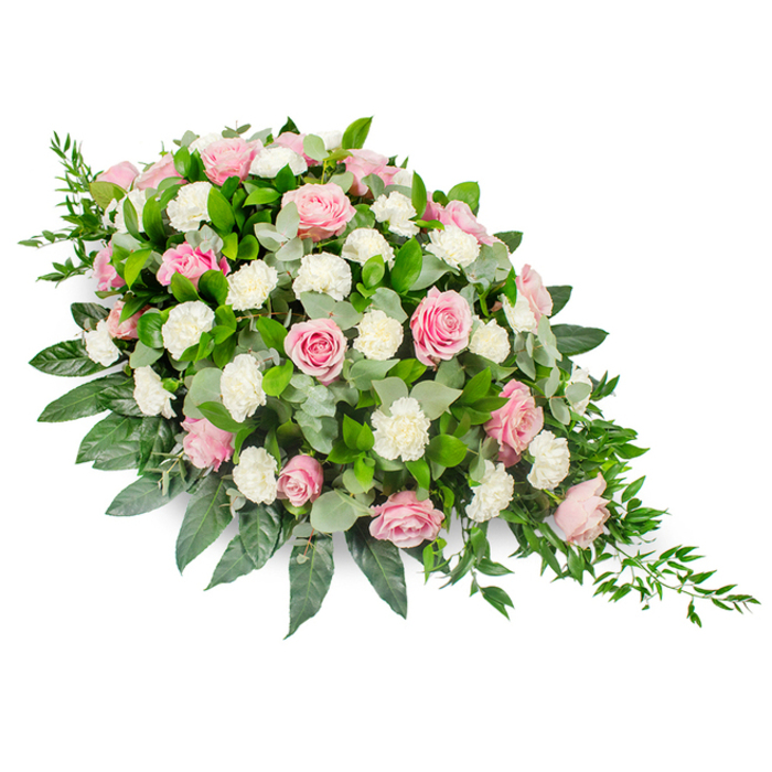Pictured against a clean white background, this substantial casket spray forms a classic, elongated teardrop, with the fullest concentration of flowers toward the upper left and a graceful tapering point toward the lower right. The arrangement is a carefully balanced blend of soft pink roses and white carnations, each element chosen to convey peace and love. The roses, in shades from delicate pastel blush to deeper rose-pink, are arranged at varying stages of openness; some remain partially furled, while others are fully bloomed, revealing intricate layers of velvety petals that seem to spiral towards a gentle centre. Between the roses, clusters of white carnations and spray carnations introduce a different texture, their finely ruffled petals catching the light and creating a plush, cloud-like appearance that softens the overall look. The entire composition rests upon a generous foundation of foliage. Glossy, dark green leaves, likely salal or a similar leaf, form a confident and stable base that outlines the teardrop shape, while rounded eucalyptus leaves in soft, silvery-green tones weave through the flowers, adding depth and a quiet, cooling note. From the edges and most notably from the narrow tip, slender sprigs of trailing greenery, possibly ruscus, cascade gently outward, adding natural movement and a hint of garden-like grace. The mood of the arrangement is tender and composed, making it an appropriate tribute for a Stanmore funeral or memorial, whether placed at a chapel near Uxbridge Road or by a graveside close to the peaceful green of Canons Park. It expresses deep condolences and remembrance through its soft colours, thoughtful shape, and expertly layered textures.