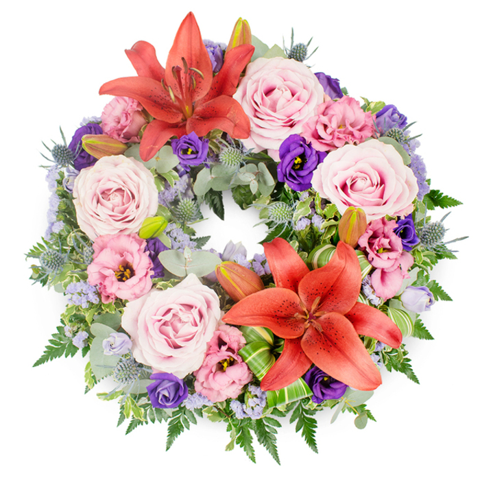 A beautifully composed circular wreath lies against a plain white background, its rich detailing revealing the care that has gone into this funeral tribute. The wreath's circular form is unbroken and full, symbolising continuity and eternal love, while the colours flow smoothly from warm coral-orange to cool lavender and deep purple. Two striking Asiatic lilies in a vivid reddish-orange shade serve as the main focal points on the ring, one near the top centre and the other slightly off-centre toward the bottom. Their wide, satiny petals curl elegantly back, and their stamens stand prominently, adding a sense of life and movement. Encircling these focal lilies are numerous fully opened roses in soft pink and lavender-pink tones, their spiral centres and gently rolled edges giving a feeling of softness and comfort. Interspersed with the roses are elegant lisianthus blooms, some in pale, almost cloud-like pink and others in luxurious, deep purple, with ruffled petals that add depth and a romantic texture. The arrangement is threaded with tiny lavender-blue flowers that resemble statice, appearing as fine, airy sprays that soften the stronger forms and lightly veil the greenery beneath. The base of the wreath is dense with foliage: rounded eucalyptus leaves in silvery green, graceful fern fronds, and darker, sturdy leaves that frame the flowers and provide contrast. Distinctive blue eryngium thistles emerge from this green base, their spiky, cone-topped heads delivering an understated contemporary accent. Closed lily buds nestle among the greenery, and carefully folded bright green leaves near one lily display skilled floral craftsmanship. This is the type of refined tribute chosen for funerals or memorials across Stanmore, whether for a quiet ceremony near Old Church Lane or a larger gathering by Bentley Priory Nature Reserve.