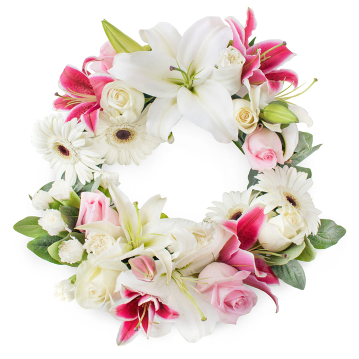 A full, open circular wreath of sympathy flowers is shown against a pure white background, the design radiating calm and quiet respect, as though ready to be placed at a memorial in Stanmore's serene surroundings. The wreath is composed of a carefully balanced mix of white, blush pink, and deep magenta blooms, each chosen to create gentle contrast and depth. Dominating the circle are large white lilies, their generously curved petals fully open and soft to the eye, showcasing central green pistils tipped with golden stamens, symbols of purity and peace. Between these, deep pink and fuchsia lilies emerge, star-shaped and sometimes speckled, adding vibrant accents that recall the rich colours of late-summer borders in local Stanmore gardens. Bright white gerbera daisies punctuate the arrangement, their layered petals surrounding dark central eyes, lending a fresh, modern note and echoing the wreath's circular form. Nestled among them are soft pink roses, in a mixture of tight buds and partially open blooms, their velvety petals bringing a tender, romantic quality and a sense of personal remembrance. Creamy white roses, fuller and more open, provide classic elegance and emotional weight, balancing the brighter lilies. Smaller white filler flowers-likely miniature carnations or spray chrysanthemums-fill in every fold of the design, contributing fluffiness and density without overwhelming the main flowers. Lush green foliage flows around and beneath the blossoms, with glossy leaves and lighter, more delicate greenery creating depth and framing the circle beautifully. Crafted with the care you'd expect from a local Stanmore florist, this circular funeral wreath is ideal for services at local churches, crematoria, or gravesides, conveying enduring love, sympathy, and honour.