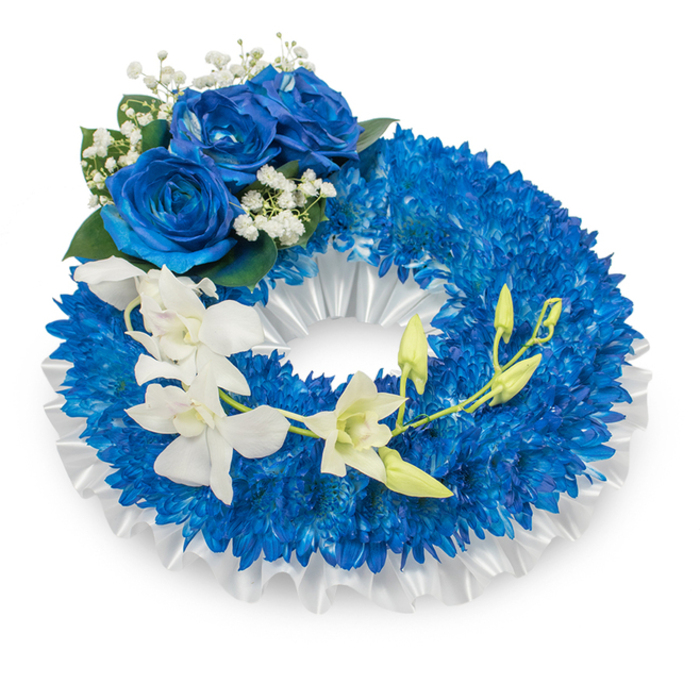 A refined funeral wreath captured against a simple white surface, this arrangement relies on saturated blue and pristine white to convey solemnity and grace. The circular form is built from a profusion of small, vibrant blue chrysanthemum-like blooms, their multitude of thin, radiating petals forming a dense, almost velvet-textured band that reads as both full and composed. A ruffled, pleated white satin ribbon nestles just inside the ring, its matte-lustrous folds providing a subtle architectural base beneath the floral mass. Prominently placed on the upper-left curve are three large blue roses, petals unfurled in a deep sapphire hue; the roses' velvety faces are complemented by fine sprays of gypsophila and modest dark green foliage, lending a classic sympathy accent. From the mid-left section a cascade of clean white orchids flows diagonally toward the lower right, the orchids' broad, delicate petals and sculpted lips creating an ethereal, drifting movement across the blue field. Interspersed among these orchids are several bright green buds - some tightly closed, some beginning to open - introducing a quiet note of renewal. The bouquet's textures and tones are rendered in soft, diffused light so every petal, ribbon fold, and leaf shadow reads with clarity. This composition is purpose-made for memorial settings - church halls, cemeteries, or quiet family gatherings in Stanmore and nearby Harrow - where its measured palette and floral craftsmanship speak of remembrance and respectful farewell.
