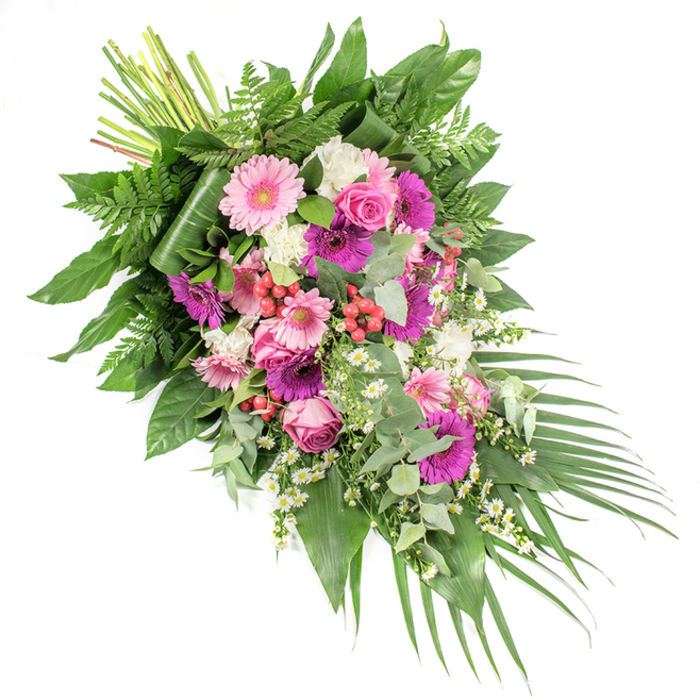 A refined, asymmetrical funeral spray is presented here on a clean white background, designed with a sweeping diagonal line that begins fuller at the upper left and gradually narrows towards the lower right. The palette combines soft pink, magenta, and fuchsia tones with serene whites and deep greens, creating a balanced, soothing tribute that would feel at home in the peaceful settings around Stanmore Country Park. At the centre of the composition, large gerbera daisies in gentle blush and more intense fuchsia hues stand out, their generous petals forming circular focal points. These are complemented by classic pink roses, each bloom carefully shaped with layered, spiralling petals that hint at enduring love and treasured memories. Between them, ruffled white flowers-resembling miniature carnations or dianthus-offer a delicate contrast, while fine sprays of small white daisy-like blossoms add an airy, whimsical texture, as if lightly scattered by a breeze. Bright red hypericum berries appear in small, rounded clusters, lending depth and a natural woodland feel. The spray is supported by a varied base of foliage: feathery green ferns, broad, glossy dark leaves that may be salal or similar, and the rounded, silvery-green discs of eucalyptus, which suggest a cool, refreshing scent. Long, sword-shaped palm fronds arch gracefully from the lower right edge, extending the line of the arrangement and enhancing its sense of gentle movement. At the upper left, the stems are neatly gathered and visible, revealing the hand-tied craftsmanship applied by a local Stanmore florist preparing this piece for funeral or memorial delivery. The overall effect is one of understated elegance, tenderness, and sincere condolence.