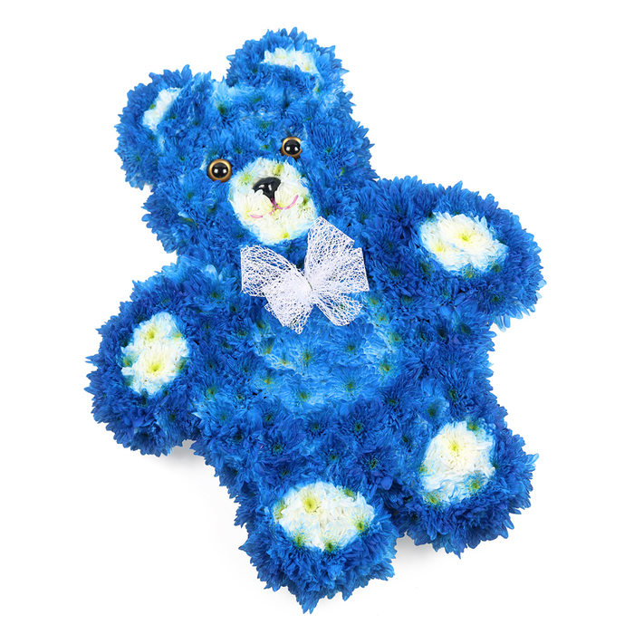 In this photograph, a teddy bear shaped entirely from fresh flowers is presented on a pure white backdrop, allowing every curve and colour to be clearly appreciated. The bear lies flat, its form slightly tilted to one side as though it has been carefully placed down. The main body, including the rounded head, tummy, arms, and legs, is densely covered with royal blue flowers, tightly packed together so that almost no gaps are visible. These diminutive blooms, similar to button chrysanthemums, give the bear a thick, plush appearance, almost like a soft fleece blanket in flower form. The deep blue tone has a soothing quality, recalling the peaceful, open skies seen from the hills around Stanmore Country Park. To bring out the bear's facial details and paws, the florist has used clusters of creamy white and pale yellow-washed flowers on the inner ear panels, the circular muzzle area, and the padded ovals on each paw. This soft contrast creates a gentle highlight, making the features instantly recognisable and lending the piece a sense of innocence. Two dark, glossy eyes gaze outward with quiet sweetness, set above a small black triangular nose. Just beneath, a simple curved line forms a subtle smile, suggesting comfort and reassurance. Around the neck, a delicate, finely woven white mesh bow is tied, its see-through loops and tails adding grace and a touch of formality, much like a special ribbon chosen for a remembrance service. The overall impression is of a tender, heartfelt floral tribute designed by a skilled Stanmore florist, ideal for child or baby boy funerals, memorials, or private commemorations. It transforms familiar teddy-bear imagery into a gentle symbol of love, memory, and ongoing care.