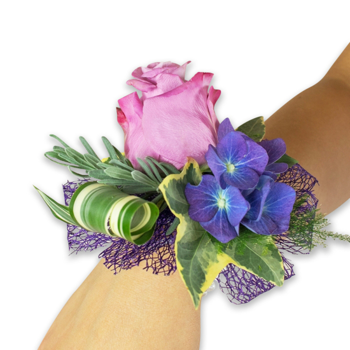 Arranged with the attention we bring to events across Stanmore, this wrist corsage is photographed on a pale forearm against a stark white background, highlighting each carefully chosen element. The centerpiece is a partially open pink rose-soft, gently cupped petals graduating from a lighter blush interior to a richer fuchsia-pink at the edges-exuding a subtle, classic rose scent that suggests early morning walks through Canons Park. Surrounding the rose are three rich clusters of deep indigo hydrangea; each tiny floret has a delicate bluish-white eye that shimmers against the darker petals and provides complex texture. A selection of slender silvery-green foliage-evoking eucalyptus or lavender-creates a soft, feathery halo behind the blooms, while glossy variegated ivy leaves edged in cream lend a touch of formal elegance. A contemporary touch is introduced with a broad, white-and-green striped leaf skillfully curled into a spiral, adding sculptural movement and a modern silhouette. The composition is seated on a layer of dark purple open-weave mesh that peeks through like evening fabric and ties the purples together. Handcrafted by florists familiar with local weddings and proms near Stanmore High Street and Bentley Priory, this corsage from Flower Delivery Stanmore reads as refined and celebratory-suitable for bridesmaids, graduates, or anyone who wants a wearable bloom that captures both tradition and a contemporary edge.