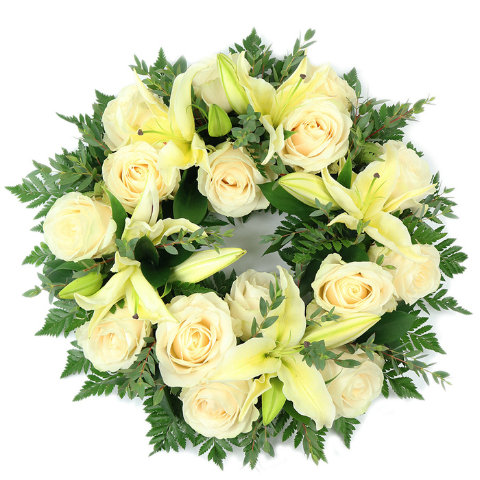 This elegant circular wreath, photographed flat on a white backdrop, balances soft cream roses and pale yellow lilies across a full, verdant base of foliage. Approximately fifteen to twenty creamy roses, each with tightly furled petals and a soft, ivory sheen, create the wreath's primary volume; their rounded forms are evenly spaced so the composition feels both abundant and ordered. Between these roses, trumpet-shaped lilies in a gentle lemon hue provide vertical sweep-some blossoms lie fully open with tapered petals and pale stamens visible, while others remain in bud, offering moments of quiet anticipation and varied texture. The supporting greenery is deliberately layered: filigree-like fern fronds soften the outer edge and lend an airy silhouette, while clusters of smaller, rounded leaves-suggestive of eucalyptus-add darker, cooler tones and a subtle aromatic note when handled. The lighting is even and soft, allowing the creams and pale yellows to glow against deep greens and the white background, emphasizing the wreath's perfect circle as a symbol of continuity and memory. As a locally based florist familiar with services at Stanmore station chapels and the green spaces around Stanmore Common and Bentley Priory, I find this arrangement both comforting and composed, suitable for funeral presentations, memorial displays, or as a heartfelt expression of sympathy that reads as both respectful and quietly hopeful.