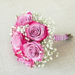 In this Stanmore-crafted bouquet, the art of floral composition is beautifully realised through colour harmony and texture. Five prominent roses, their petals shifting from soft, muted lavender at the centre to vivid magenta along the edges, bask in gentle daylight-a visual echo of Stanmore's quiet, leafy avenues. Wisps of white gypsophila (baby's breath) drift among the blooms, imparting a cloud-like volume and contrast that speaks to both innocence and grace. There's a tactile richness: robust, layered rose petals lie alongside the airy fineness of baby's breath, all gathered and finished with a dark purple satin ribbon. Iridescent pearls spiral neatly around the handle, lending a finishing touch of timeless luxury. The bouquet rests on a pale oatmeal linen reminiscent of the subtle textures found in Stanmore's family homes and event venues. Every element, from the exposed green stems to the choice of elegant wrapping, reflects the thoughtful attention of a Stanmore florist. The arrangement emanates warmth, artistry, and joyful elegance, making it a perfect fit for intimate occasions, wedding ceremonies, or special local celebrations.