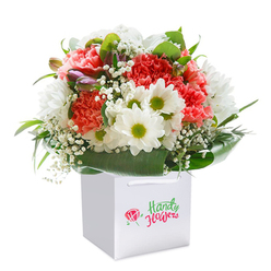 Echoing the welcoming ambiance of Stanmore, this stunning floral arrangement is a study in contrasts and harmony. Generous coral and peach carnations, each petal ruffled and vibrant, anchor the bouquet with warmth and charm, recalling walks along Stanmore Hill adorned by spring blossoms. Spots of vivid red bring depth, while bright white daisies with cheerful yellow-green centers provide a fresh, lively accent. Tiny clouds of white baby's breath add delicacy, softening the edges with their airy presence. On one side, hints of purple-tinged buds emerge, suggesting growth and anticipation-much like the ever-changing beauty of Canons Park through the seasons. Glossy, deep green leaves encircle the entire design, their rich foliage forming an elegant natural backdrop. Nestled inside a modern white gift bag with sturdy ribbon handles, the arrangement is made for effortless celebrations and memorable deliveries throughout Stanmore. The pristine white background ensures every nuance is captured-each stem arranged with intention and pride by experienced local florists, making this bouquet ideal for birthdays, anniversaries, or to simply brighten a loved one's day in Stanmore.